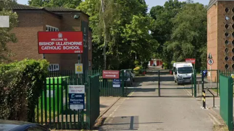 A large brick building and rows of trees can be seen behind green gates. A white van and cars are parked outside. A large red sign with Welcome to Bishop Ullathorne Catholic School can be seen hanging at the front of the site.