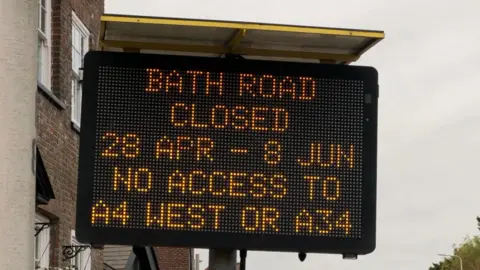 A digital road sign has been lit-up to read "Bath Road closed 28 Apr - 8 Jun. No access to A4 west or A34".  Houses can be seen behind the sign and grey skies overhead.