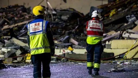 Government of Jersey Two firefighters are standing in front of a large pile of rubbish on the site of the explosion. They are in uniform and are wearing hard hats. The man standing closest to the camera has FIRE SAFETY OFFICER printed on the back of his jacket.