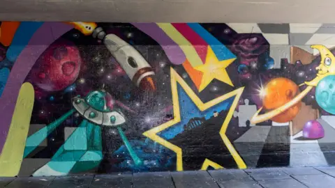 Newcastle City Council A mural in one of the underpasses featuring an alien spaceship, a rocket, planets and the Tyne Bridge within a yellow star.