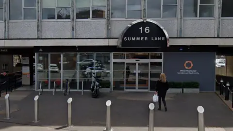 Google The outside of a building with West Midlands Combined Authority sign outside, with the address 16 Summer Lane written in white on a black sign above a glazed door. A woman with long blonde hair walks towards the entrance, which is behind silver bollards and has a moped parked against bike racks outside. Two stories of the building can be seen: the lower level is fully glazed, with windows along the upper level.