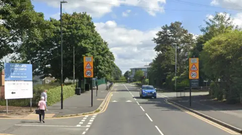 Google A sign saying 'Failsworth Sports Campus' is at the side of a road. School 20mph signs are either side of a road. One older person is walking on the pavement and another older person is crossing the road.