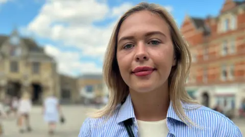 Daisy Blakemore-Creedon has blonde hair and is standing outside in Peterborough city centre in front of the Guildhall building. She is wearing a butter yellow coloured T-shirt with a white and blue striped shirt over the top. She is looking directly at the camera and the sky is blue in the background behind her.