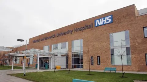 PA Media The brick exterior of the entrance to the Norfolk and Norwich Hospital. There is a large sign on the front and a partially covered glass awning in front of the door.