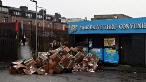 A large pile of more than 50 cardboard boxes dumped in the corner of a car park. It is next to a shop, the sign reads: 24hr drive thru convenience
