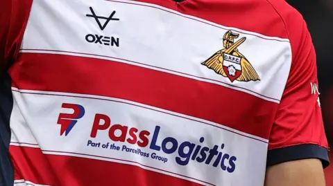 A close up of a Doncaster Rovers football kit. The shirt has read and white hoops and a Doncaster Rovers badge on it. The kit is sponsored by Pass Logistics.