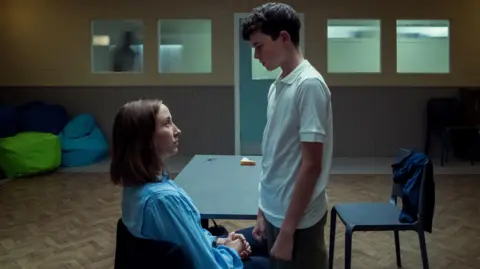 Netflix Erin Doherty and Owen Cooper in Adolescence. Erin is sitting down in a blue shirt or blouse. Owen is standing up, looming over her, in a white polo shirt, his fists clenched. They are in a large empty room, with a table and chairs.