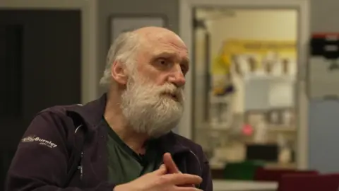 Chris Keene talks to a BBC reporter, situated off camera. He has white/grey hair and a white beard. He is wearing a navy jacket and green-tshirt and sits in a community centre.