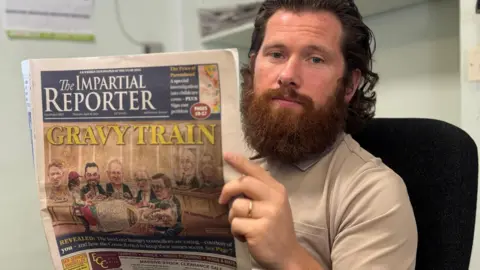 Ciarán Flaherty, news editor at the Impartial Reporter, holding up a copy of the Impartial Reporter with the headline 'Gravy Train' and an cartoon depiction of inside the council chamber. 