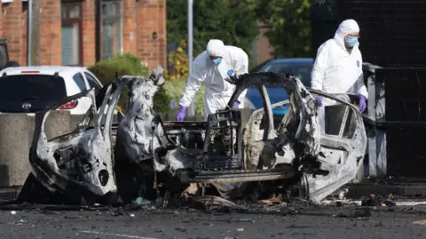 Pacemaker Press Two forensic officers are next to the remains of a car that's exploded. Parts of the frames remain. Debris is lying around on the street. The forensic officers are wearing white overalls and a blue mask on their faces.
