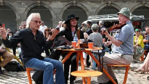 Damien Eagers/PA Wire Three men sit at a table in the foreground of the picture. One man, to the right of the frame is playing a violin. He is wearing a light blue shirt, cream trousers and a green hat. He is facing towards the table. In teh middle of the table is a man with long dark hair and a short beard. He is wearing a black jacket and the strap of the guitar, he is playing, is visible. A third man faces towards the left hand side of the table. He is wearing a dark shirt and blue jeans. He has curly blonde or grey hair and sun glasses. He is holding a brown case in his hand. Other people are sitting in the background, listening to the music.