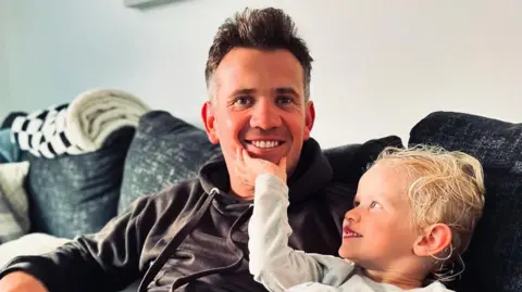 Andy Johnson Photograph of Andy Johnson (left) pictured with his son, Oaklen. His son looks up at him and holds his chin in his left hand, while Andy smiles at the camera. 