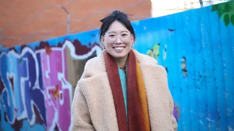 Louise Ndibwirende Grace wearing a thick winter coat and scarf, standing smiling in front of a fence with brightly-coloured street art