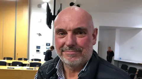 A picture of Dave Taylor. He is facing the camera and smiling. He is standing in a council room with tables and chairs behind him. He is bald and has a grey beard. He is wearing a blue gilet jacket over the top of a striped blue and white shirt.