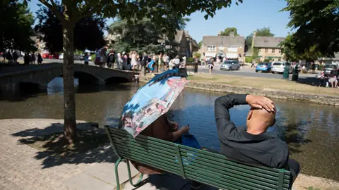 Getty Images Two people sitting on a green metal bench beside the river in Bourton on the Water. It is a warm sunny day and one of them is sitting under the shade of a parasol. 