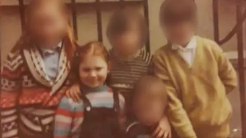 Karen McLaughlin A group of five young children are huddled together in front of black metal railings outside a building. They are all in brightly dressed clothes. Four of the children's faces are blurred, but one young girl - in a striped tshirt and dungarees, is seen smiling at the camera, with her eyes closed as she faces the sunlight. She has ginger hair and one of her arms wrapped across the shoulders of a smaller boy stood next to her. 