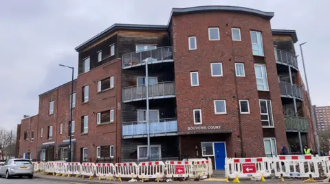 An exterior shot of Bouverie Court, it is a red brick building with a mix of terraced housing to the left and a three-story block of flats with large balconies on the right hand side of the building. There are roadworks going on on the pavement outside