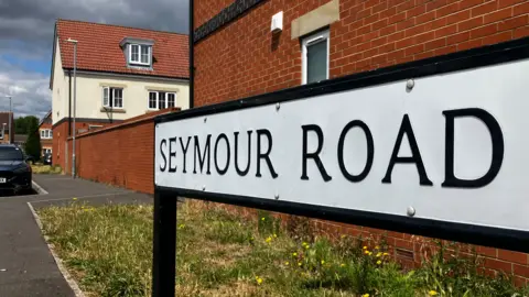 BBC A close up image of a sign reading "Seymour Road" with scrubby grass, a car and red brick buildings in the background.