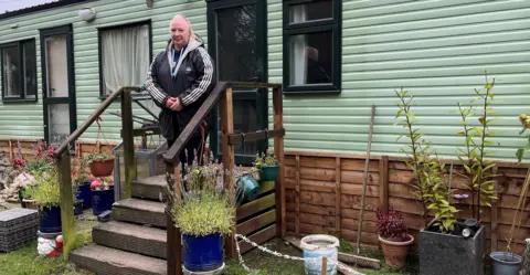 A woman on the steps of her green static caravan in the Borders surrounded by various plants