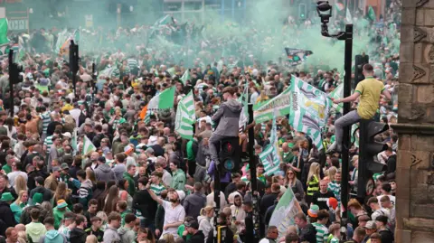 PA Media A large crowd of Celtic supporters gathers in a Glasgow street, with green smoke in the air and flags being waved, while some people sit on traffic lights.