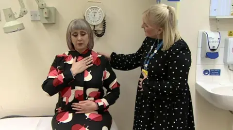 A patient being treated at the long Covid clinic. She has a brightly patterned shirt on and is sat on a bed with her hand on her chest while a physiotherapist stands next to her and is testing her breathing. 