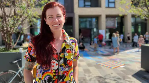 Kaye stands outdoors in front of a modern building, wearing a colorful shirt with rainbows, hearts, and the word 'HAPPY' repeated across it. The paved area around them is decorated with chalk drawings, and people are walking or sitting nearby. Trees and a bicycle are visible to the left