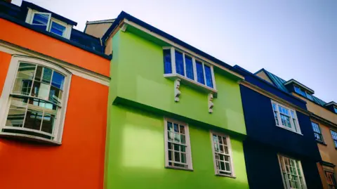 Elsie from the block Three houses on a street. One is bright orange, one is bright green and one is navy blue. They all have white windows with reflections of the street and the sky overheard is blue