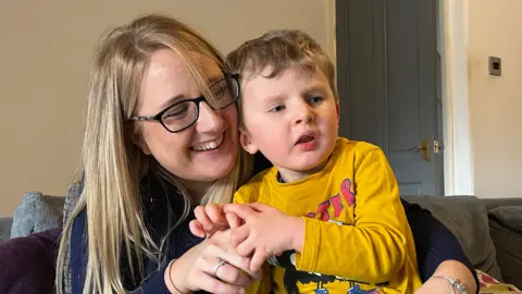 A mother and her son sat on the sofa. The mother is wearing glasses and holding her son in her arms. He is looking away from the camera.