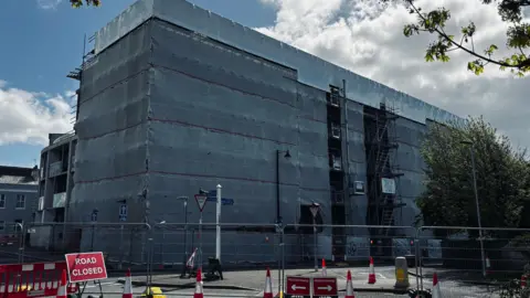 This image shows a large multi‑storey building completely wrapped in grey protective sheeting, with scaffolding visible along one side. The covering stretches from ground level to the roof. Several layers of scaffolding are exposed on the right‑hand section, with metal poles, platforms and ladders visible through gaps in the sheeting.