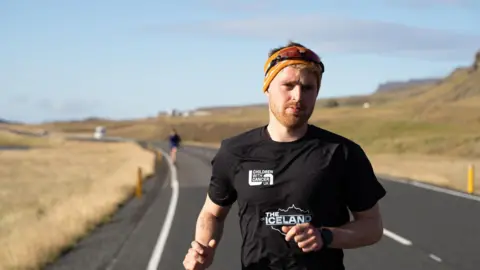  Robbie Guthrie/PA Seb Key running in Iceland - he is wearing a black t-shirt and a headband. He is running on a road, with yellow-ish fields each side.