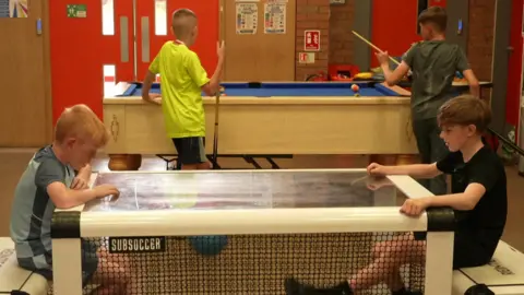 BBC Two young boys are playing a football game in a cage with two other boys playing pool behind them. They are all in the main hall of a youth club.