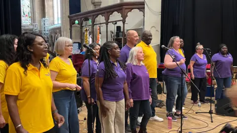 Thirteen members of a choir standing on a stage behind a number of mics. They are stood in a church like hall.