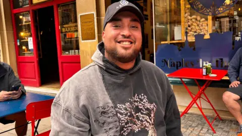 A smiling man in a baseball cap sits on a terrace outside a cafe