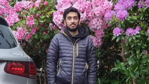 Family A man stands beside a parked car in a garden filled with bright pink and purple flowers. He is wearing a padded jacket and trousers, positioned in front of a red brick house.