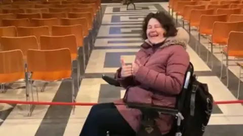 BBC A woman in a light red coat smiling and sticking up her thumbs in a hall surrounded by wooden chairs