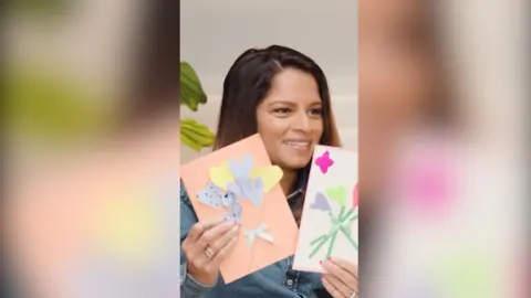 A woman in a jean jacket holds up two greetings cards, made by children.