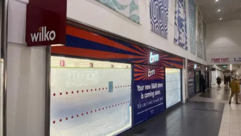 Andrew Turner/BBC Wilko branded sign adorns the shopping centre facade with new B&M Bargains signage now showing, with polythene screens in the shop window to obscure refitting work inside. To the right of the image is the shopping centre, featuring grey and white tiled floor, and abstract design panels above the shop facade.