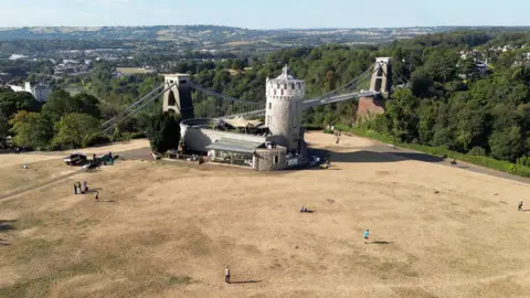 An aerial view shows Clifton Observatory next to Clifton suspension bridge west Bristol, with parched parkland around it with people walking on the brown grass, on 9 August.