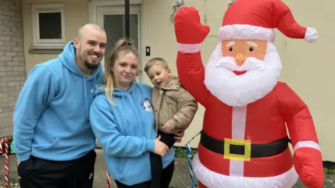 Elias in his mum's arms, wearing a dark cream jacket and black trouser. His mum and dad are in blue jumpers with matching logos. There is a big inflatable Santa next to them all, outside their home in Radstock.