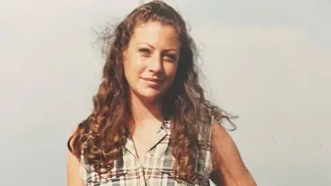 A photo of a young woman with long auburn curly hair wearing a pale checked sleeveless top