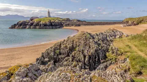 Getty Images Image shows rocky terrain and a sandy beach in the foreground.
A small island in the background, topped with a lighthouse and an adjacent building.
The island is surrounded by the sea, with mountains visible in the distance.
The sky is mix of blue sky and some clouds above the mountain's on the horizon.