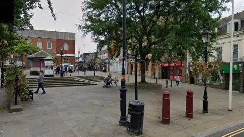 Google A paved area in Wellingborough town centre, with shops to the left and right. There are trees on the edge of the paved area, and a set of steps leading up to a small self-contained pagoda-shaped building. There are a few pedestrians walking around.