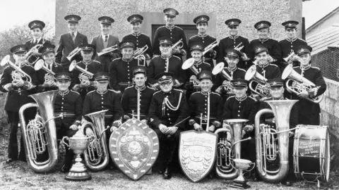 A black and white photo of Indian Queens Band assembled in front of their bandroom with several trophies and their instruments
