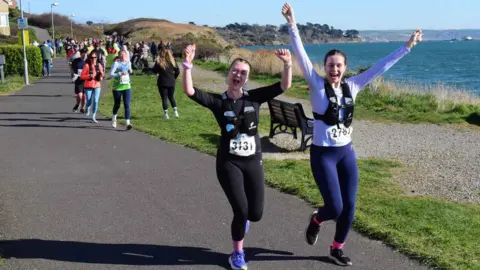Two women wearing running gear and race numbers raise their hands in the air and cheer as they run past the photographer on the seafront