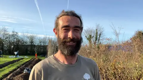 Ben Parkes, stood in a field, with work going on behind him. He has a black beard and black hair. He is wearing a brown t-shirt and is smiling. He is surrounded by bushes and trees.