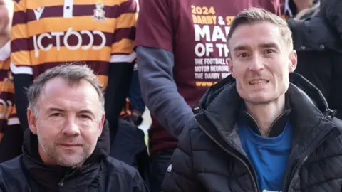 PA Media Marcus Stewart and Stephen Darby smile at the camera. Both have short dark hair and wear black coats. 