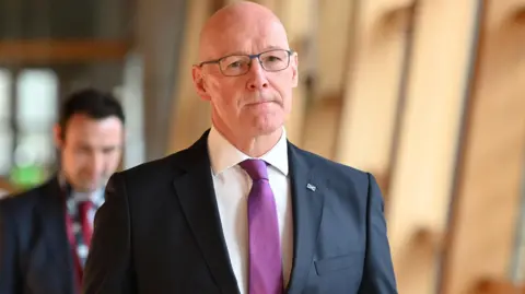O primeiro ministro da Escócia, John Swinn, caminha no parlamento escocês, usava um terno escuro, camisa branca e gravata roxa.