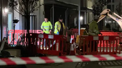 BBC A picture of workers outside of a property in Jersey attempting to identify the gas leak. They are surrounded by a cordon of fences and tape.
