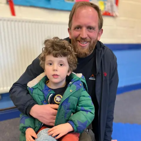 Emily Doughty / BBC Dad Sam sits on a padded floor holding a small child wearing a green coat and red trousers. The child sits on his dad's lap with a hat resting on his legs. They are indoors against a painted brick wall with colourful displays in the background.