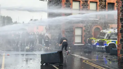 Pacemaker A group of people standing watching as someone in a tracksuit runs across a spray of water canon in front of a residential street and two police vehicles.